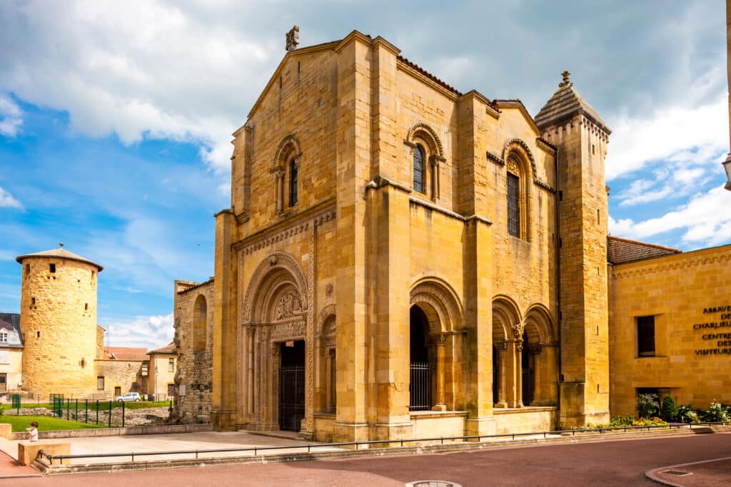Abbaye de Charlieu : Splendeur de l'église romane Abbaye romane de Charlieu en pierre jaune, avec une tour ronde et l'entrée sculptée. Panneau 'CENTRE DES VISITEURS'. Ciel nuageux.
