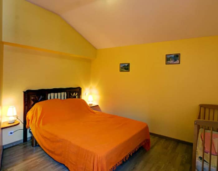 Chambre mansardée lumineuse aux murs jaunes, avec un lit double orange, tables de chevet et lampes. Un berceau en bois est présent.