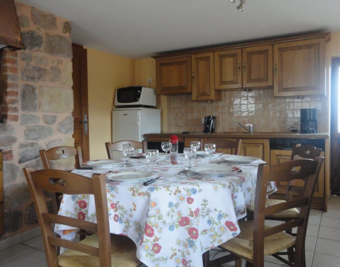 Table ronde dressée avec nappe fleurie et chaises en bois dans une cuisine rustique avec mur en pierre et mobilier bois.