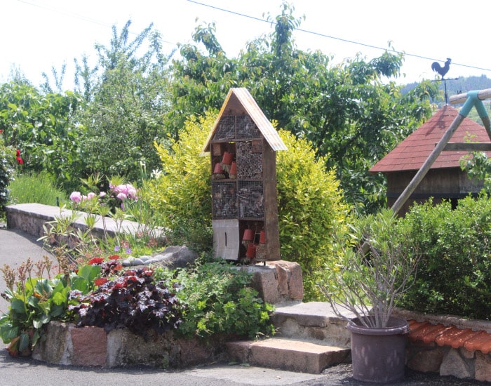 Hôtel à insectes et jardin : oasis fleurie pour la nature Grand hôtel à insectes en bois dans un jardin verdoyant et fleuri. Des pivoines roses et un coq sur une girouette sont visibles.