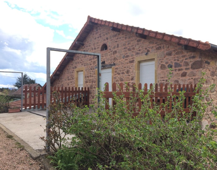 Maison en pierre avec toit de tuiles, fenêtres à volets blancs, bordée par une clôture en bois et des buissons verts le long d'un chemin.