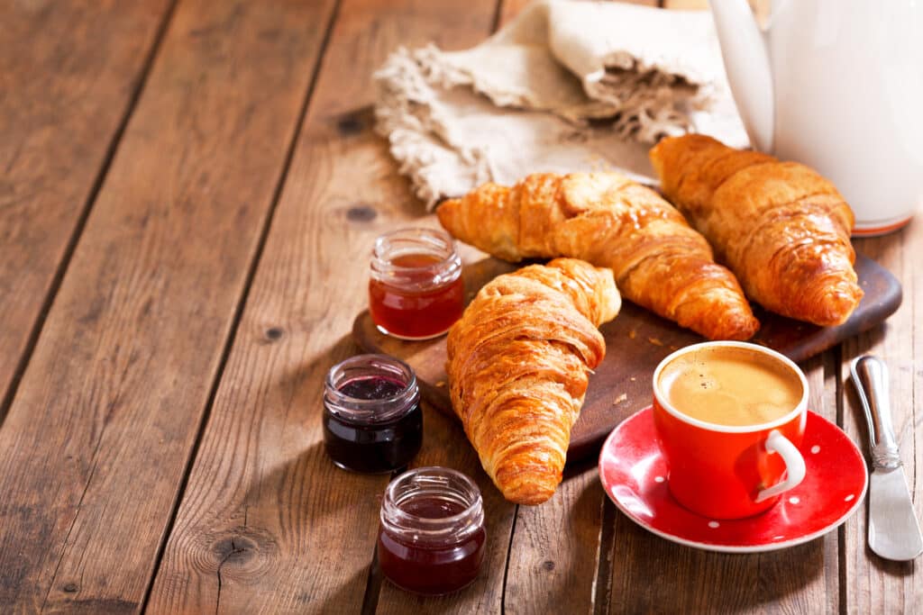 Trois croissants dorés, une tasse de café rouge, trois pots de confiture sur une table en bois rustique pour un petit déjeuner.